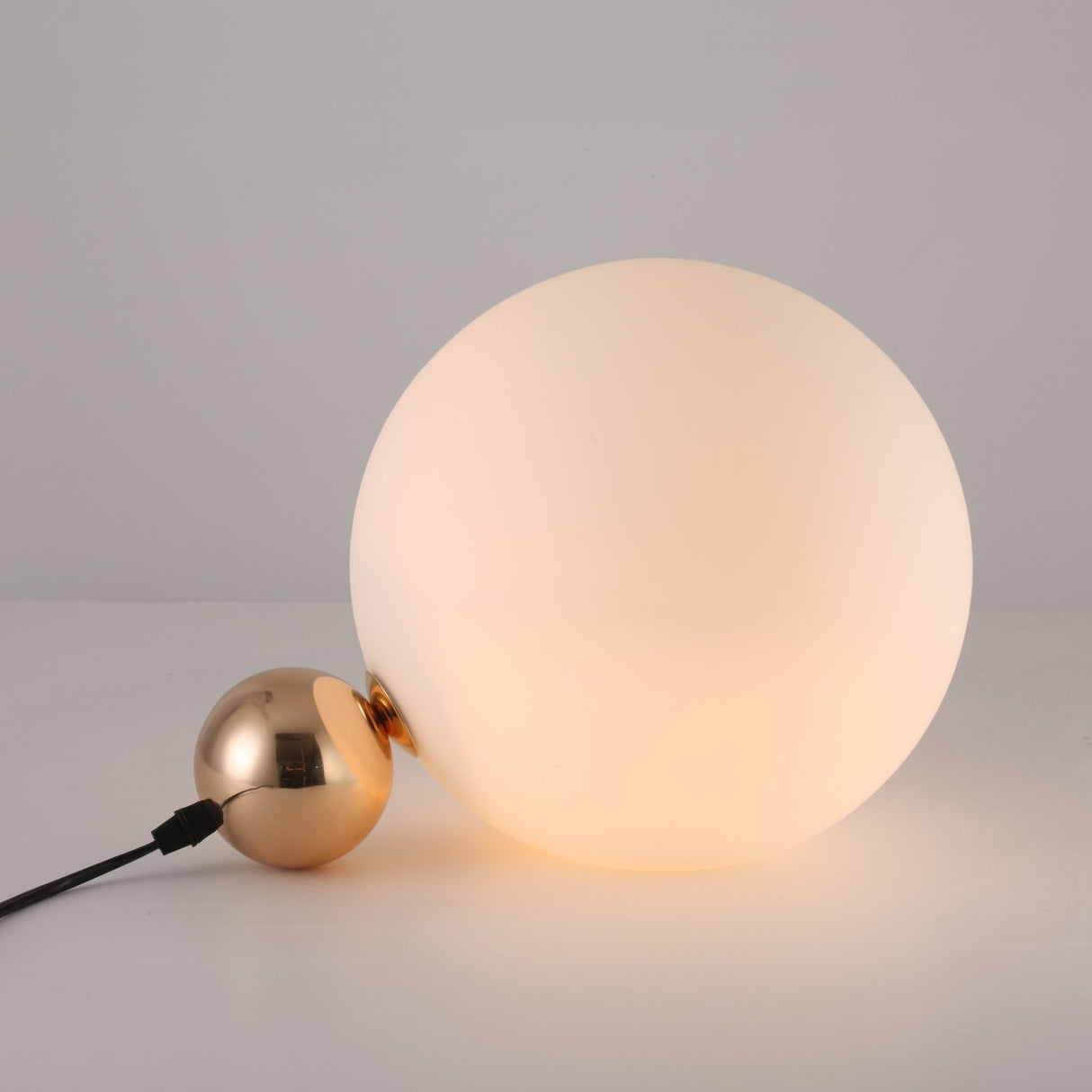 The Soul Table Lamp - Gold is a modern desk lamp with a large frosted white globe atop a small metallic gold sphere base, connected by a black power cord and set on a plain white surface.