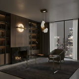 A modern office with a glass desk, armchair, built-in bookshelves, a marble fireplace, and large windows overlooking a city skyline at night—illuminated by the Caelis 4 Light LED Alabaster Cluster Pendant Light in brushed brass and soft white.