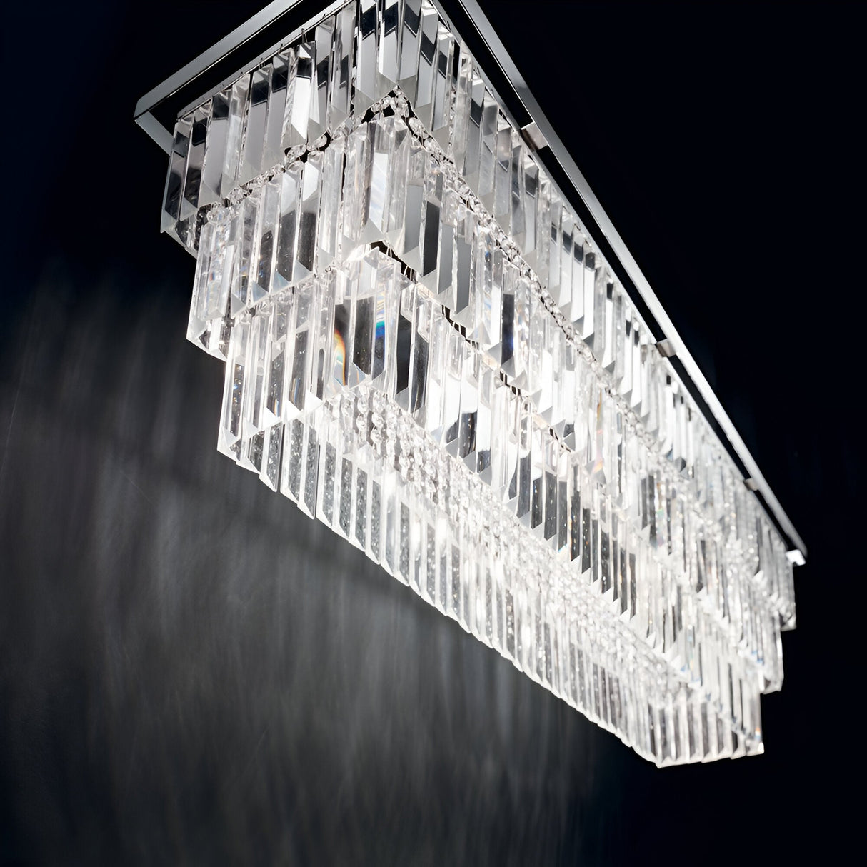 The Fluxon 6 Light Crystal Suspension - Chrome, a modern rectangular chandelier with multiple tiers of glass prisms, is illuminated, casting bright reflections against a dark background.