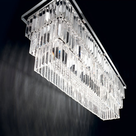The Fluxon 8 Light Crystal Suspension - Chrome is a striking rectangular chandelier that stands out against a dark background. Its contemporary style showcases multiple layers of hanging prisms, which capture and scatter light to produce a dazzling shimmering effect.