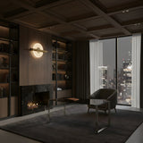 A modern office with dark wood bookshelves, black marble fireplace, glass desk, and gray armchair. Large windows reveal a city skyline at night. The Elyra LED Alabaster Wall Light in brushed brass adds warmth and sophistication.