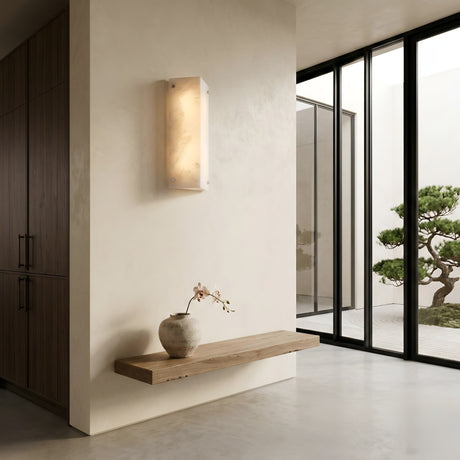 A minimalist interior features a floating wooden shelf with a ceramic vase holding an orchid. The Auralis 1 Light Alabaster Wall Light in Soft White casts a gentle glow, while floor-to-ceiling windows overlook a manicured courtyard tree.