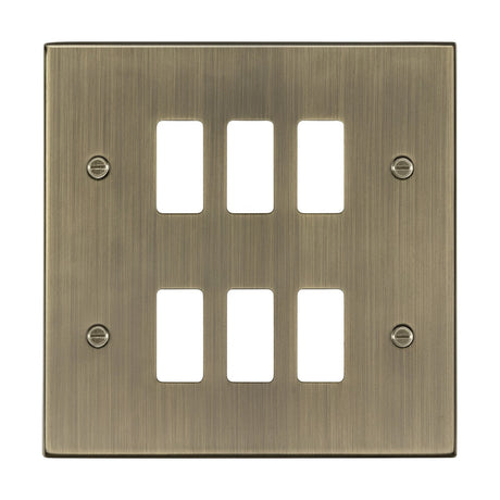 The 6 Gang Grid Faceplate in Antique Brass features a square-edged design with six rectangular slots for switches. It has a brushed finish and is complemented by four mounting screws in each corner, with the slots arranged in two vertical columns.