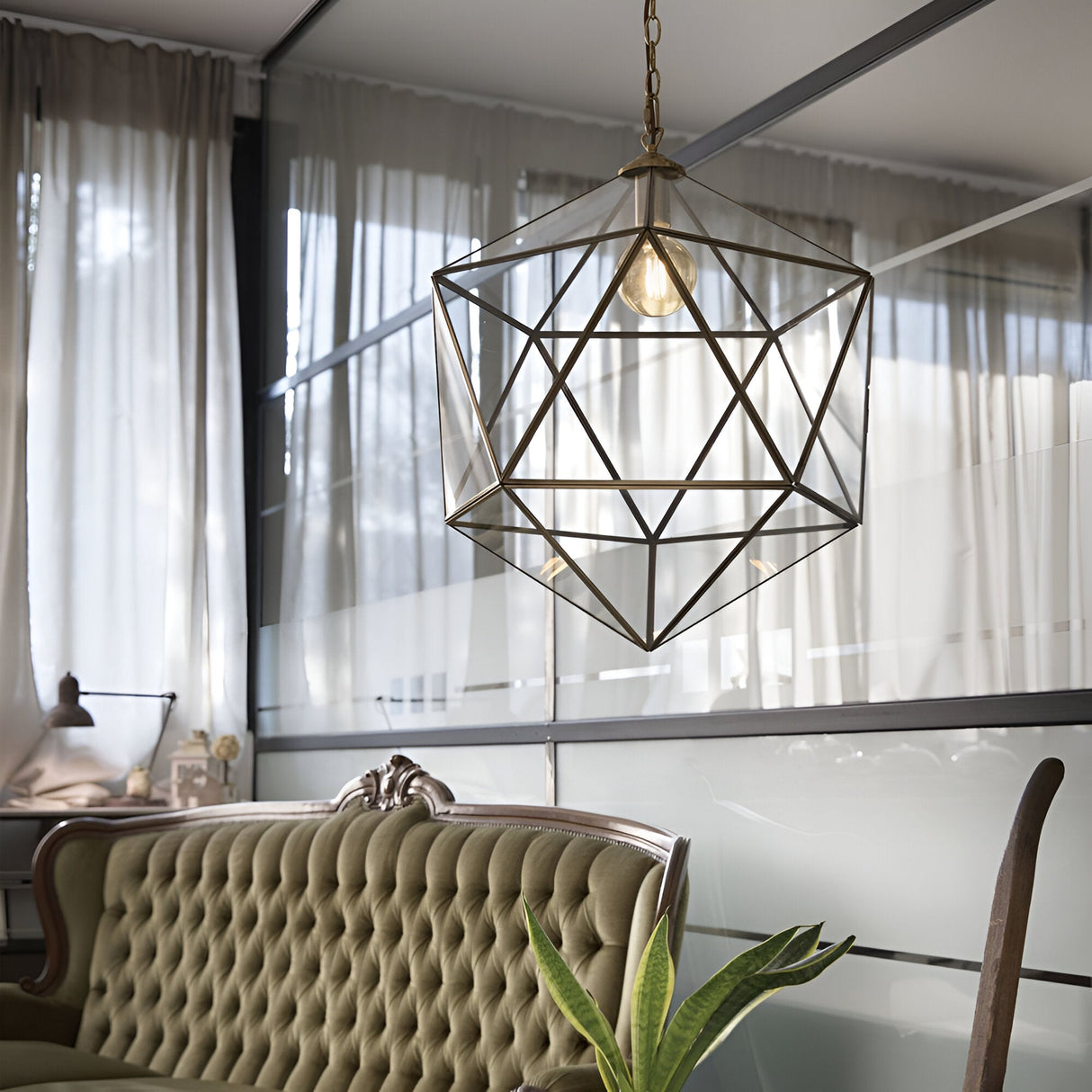 The stylish room features the Verdant 1 Light Large Pendant in a brass finish, hanging from the ceiling amidst frosted glass panels. Positioned below is a green tufted sofa, complemented by a plant placed to the right. Soft natural light gently filters through sheer curtains that cover the expansive windows.