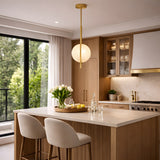 A modern kitchen with light wood cabinets, a marble island, two beige stools, glassware, large windows with green views, and an Elyra LED Alabaster Single Pendant Light in brushed brass & soft white hanging above a tray with flowers and lemons.
