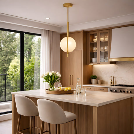A modern kitchen with light wood cabinets, a marble island, two beige stools, glassware, large windows with green views, and an Elyra LED Alabaster Single Pendant Light in brushed brass & soft white hanging above a tray with flowers and lemons.