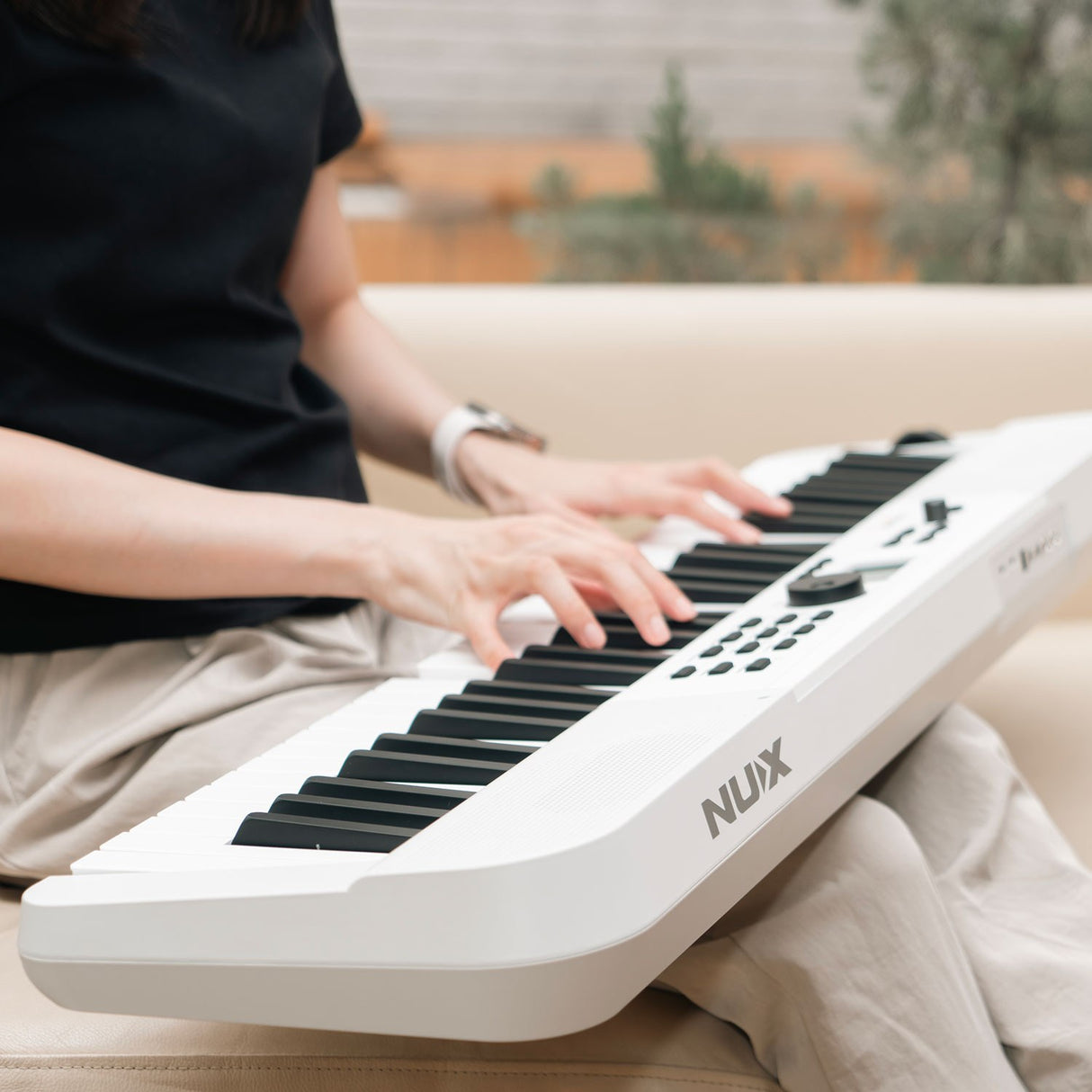 Woman in a black top playing the NU-X NEK-110 61-key portable keyboard in white, resting on her lap with control panel and pitch wheel visible.
