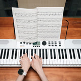 Overhead view of hands playing the AVSL NU-X NEK-110 61-key portable keyboard in white, with open sheet music on the stand above the controls.