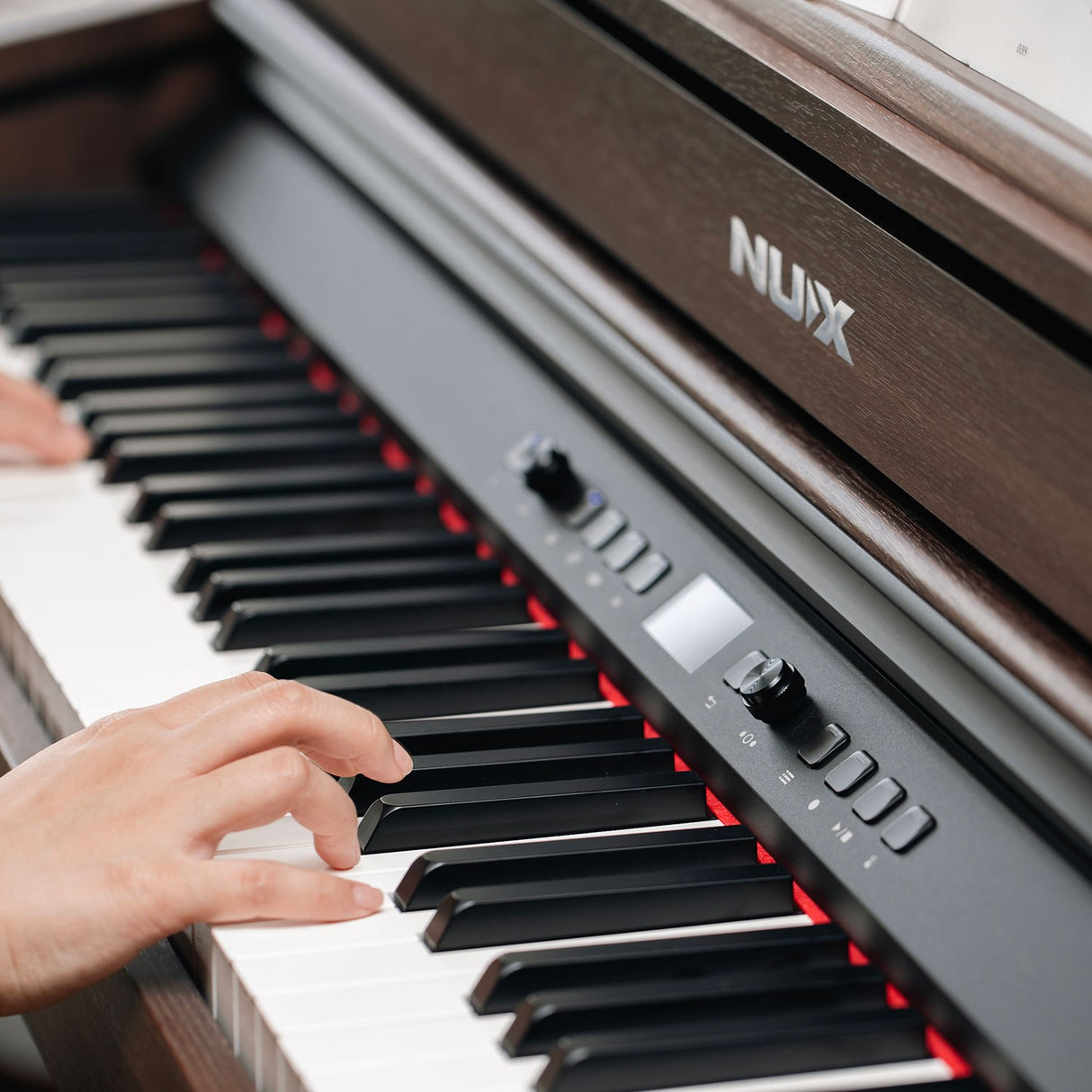 Close-up of hands playing the NU-X NCK-430 weighted digital piano in walnut, showing the black control panel with display, dial and buttons beside the keys.