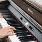 Close-up of hands playing the NU-X NCK-430 weighted digital piano in walnut, showing the black control panel with display, dial and buttons beside the keys.