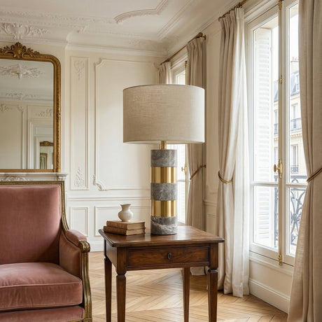 A stylish living room features a pink velvet armchair, a wooden side table with books, the Arvella 1 Light Striped Marble Table Lamp in grey and gold, tall windows with beige curtains, ornate white walls, and a large gold-framed mirror.