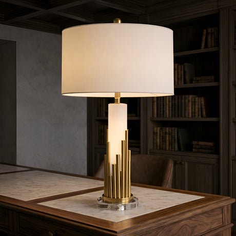 The Celora 1 Light Vertical Spire Alabaster Table Lamp in Brass & Soft White features a modern, sculptural design with a cylindrical shade, perfect for illuminating marble-topped desks or studies with bookshelves in the background.