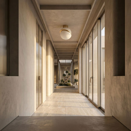 A modern, sunlit hallway features large glass doors on both sides, beige walls, and an Arc Semi-Flush Ceiling Light - Gold & White. At the end, there's a minimalist indoor garden with potted plants and gravel.