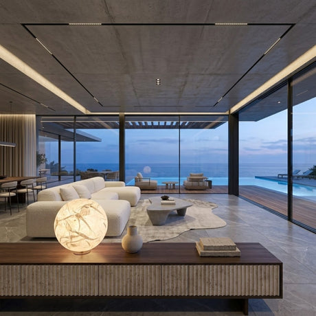 Modern living room with large glass windows, neutral-toned furniture, a glowing Lunar Table Lamp - White on a table, and an ocean view with pool outside at sunset.