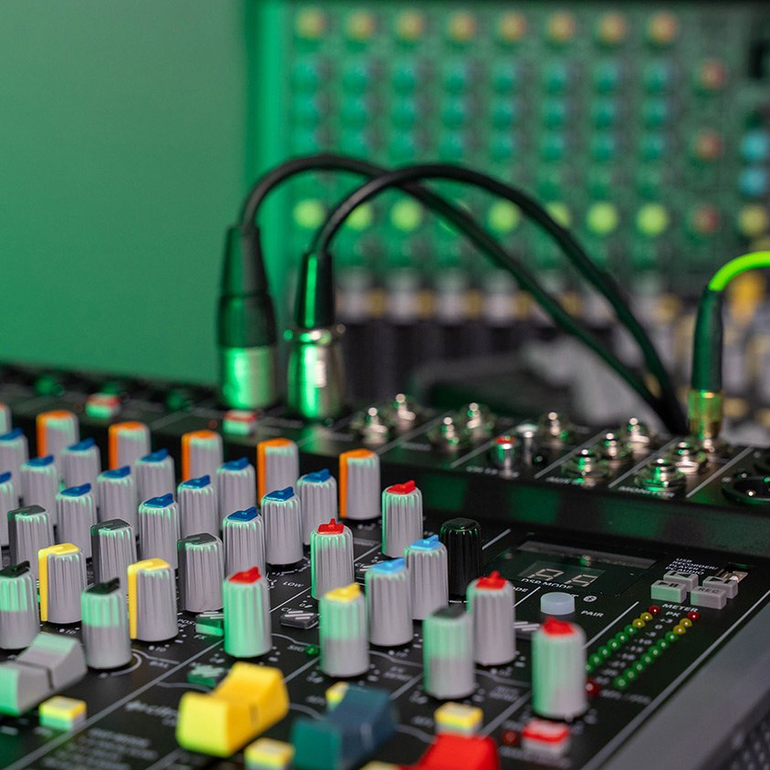 Close-up of the AVSL Citronic CMC-16 live mixing console showing coloured knobs, faders, XLR cables and illuminated controls against a green LED-lit backdrop.