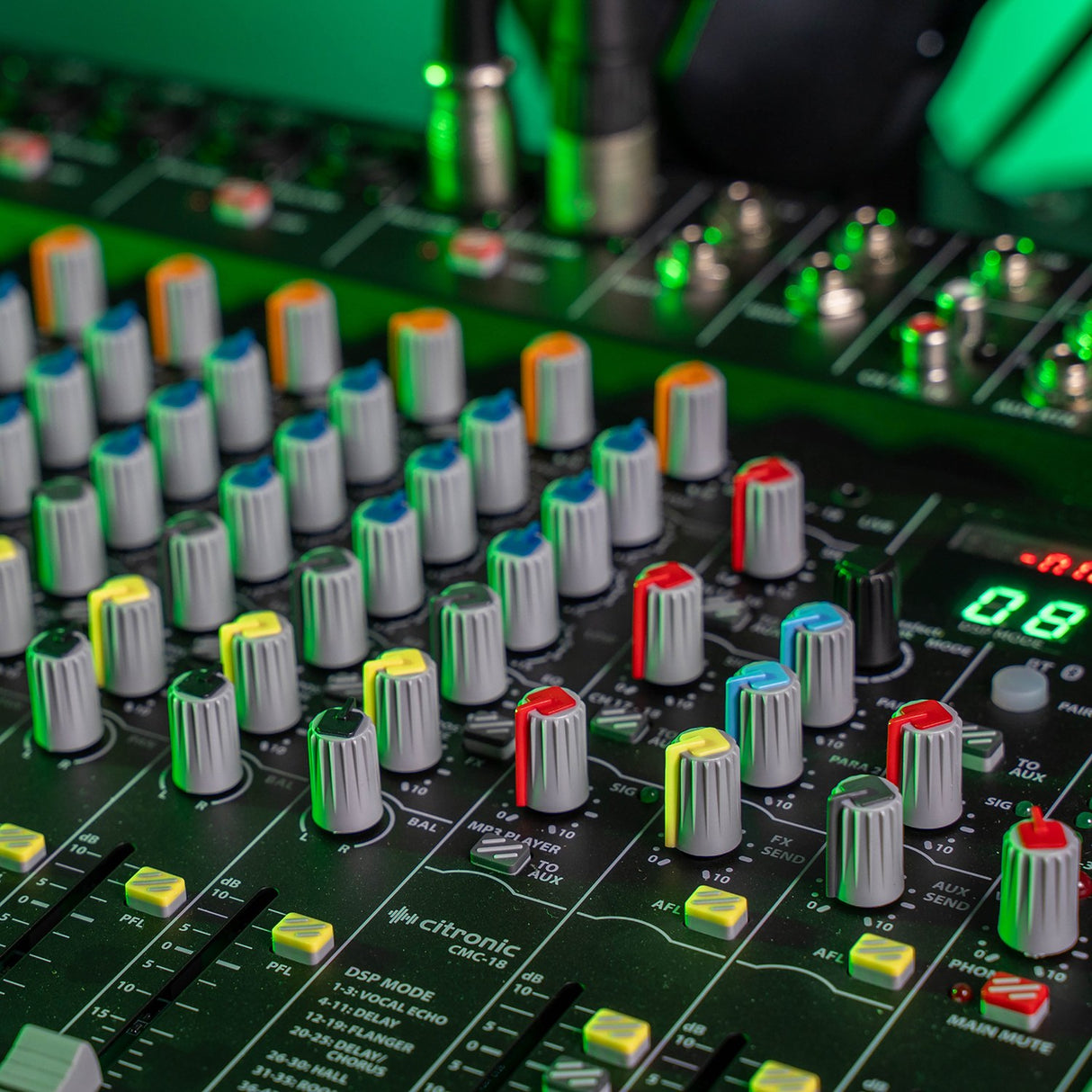 Close-up of the Citronic CMC-18 mixing console showing coloured EQ, gain and aux knobs, yellow PFL buttons, faders and DSP display under green lighting.