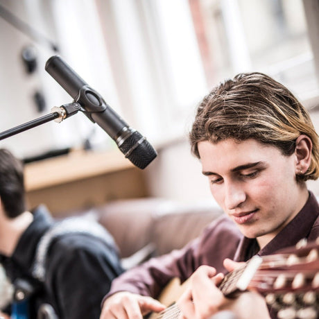 Young musician playing an acoustic guitar while singing into an AVSL Chord NUHH-864.1 handheld wireless microphone on a boom stand indoors.