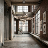 Industrial-style interior with exposed brick, large windows, and concrete floors. The Stratiform 20 Light Metal Chain Veil Chandelier in Nickel adds a modern touch. Minimalist benches line the wall, accented by potted plants and framed art.