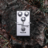 NU-X Steel Singer analog drive guitar pedal in silver with Volume, Gain and Filter knobs and foot switch, shown on a weathered tree stump.
