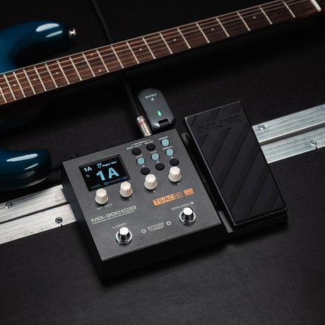 NU-X MG-300 mkII guitar multi-FX pedal with looper by AVSL, shown with expression pedal and wireless receiver beside a blue electric guitar.