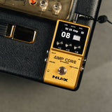 NU-X NCA-1 Amp Core Studio pedal in gold finish with colour display showing "92 US Amp" preset, three rotary controls and footswitch, beside an amplifier.