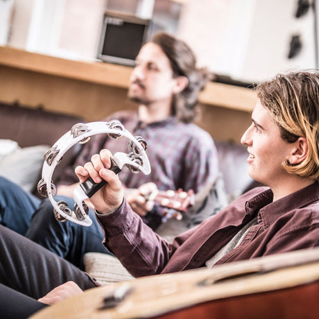Young man holding the Chord TAMB-SD-WH white D-shape plastic tambourine with paired jingles, seated on a sofa beside a guitarist in a relaxed setting.