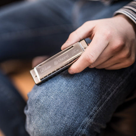 Hand holding the Chord Blues Ten diatonic harmonica in A, showing its steel cover plates with numbered holes 1–10, resting against denim jeans.