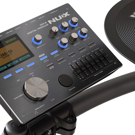 Close-up of the NU-X DM-8 digital drum module with blue-lit rotary knobs, faders, genre dial, colour display, and a black mesh cymbal pad alongside.