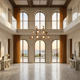 A grand, modern interior with tall arched windows and marble floors features the Petal 24 Light Chandelier in Copper, golden curtains, minimalist decor, and offers ocean and hill views at sunset.