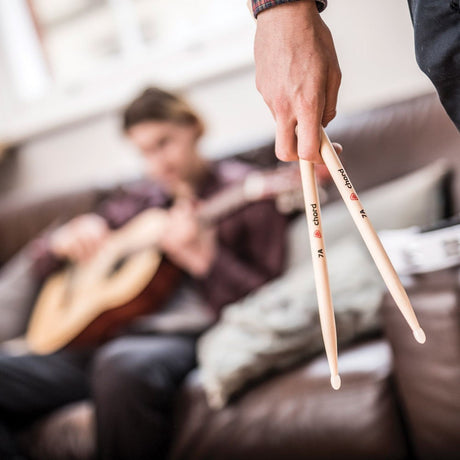 Hand holding a matched pair of Chord M7AN maple 7AN drum sticks with a guitarist seated on a brown leather sofa blurred in the background.