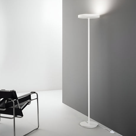 A Nobe Floor Lamp in white casts light onto a gray wall, enhancing the minimalist design. A black leather chair with a chrome frame sits adjacent, embodying modern sophistication. The scene exudes a clean, contemporary aesthetic.