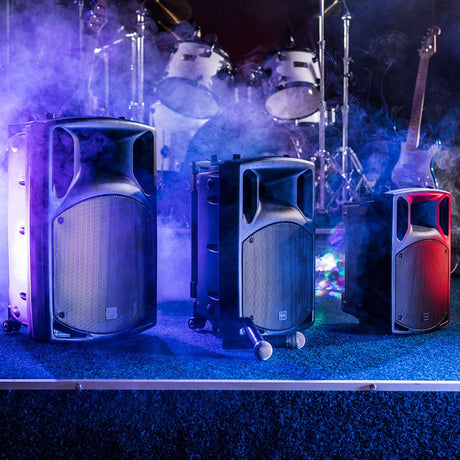 QTX QX12PA portable powered PA speakers by AVSL displayed on a smoky stage with wireless microphones, drum kit and guitar in blue and red lighting.