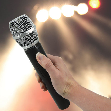 Hand holding the QTX QHH-863.8 UHF handheld wireless microphone with black body and silver mesh head, against a blurred stage lighting backdrop.