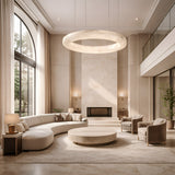 A spacious, modern living room with tall windows, neutral tones, a curved sofa, two armchairs, a round coffee table, a fireplace, plants by the windows, and the Aurexis LED Large Floating Ring Alabaster Suspension Light in soft white as a chandelier centerpiece.