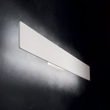 The Flara LED Wall Light 23W 3000K in white, measuring 53cm, is mounted on a dark textured wall. Its sleek rectangular design emits a soft glow, providing an energy-efficient lighting solution that offers gentle illumination around it and showcases its smooth and minimalist aesthetic.