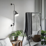 A minimalist room showcases the Etera Wall Light in black, casting a soft glow over a small wooden stool adorned with a fern. Nearby, a black clothes rack displays a patterned throw and a white shirt, and a woven basket sits close by amidst green plants beside the window.