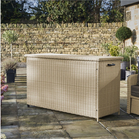 Sandy Beige Large Cushion Box K/D on a patio, a wide rectangular garden storage box in synthetic woven beige finish with hinged lid, castors and side handle.