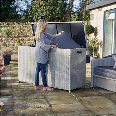Woman placing a grey cushion into the Stone Grey Large Cushion Box, a synthetic weave garden storage box on wheels, set on a patio.