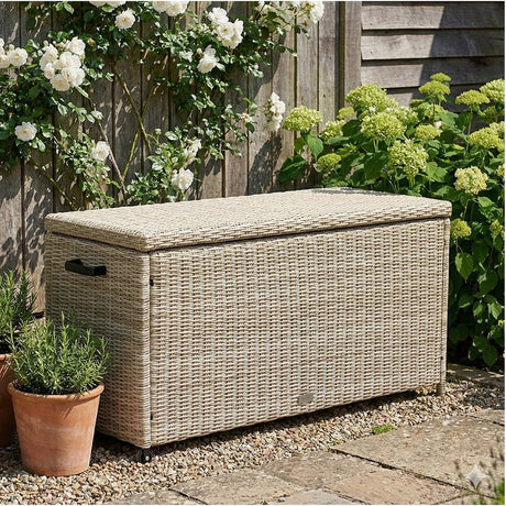 Sandy Beige Small Cushion Box, a rectangular garden storage box with hinged lid, woven synthetic rattan in beige, castors, and side handle, shown outdoors on gravel near plants.
