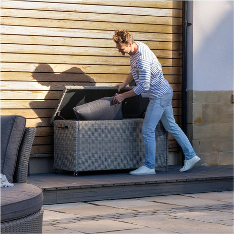 Man lifting the hinged lid of a Slate Grey Small Cushion Box, 64cm, in synthetic weave, to place a grey cushion inside on an outdoor decking area.