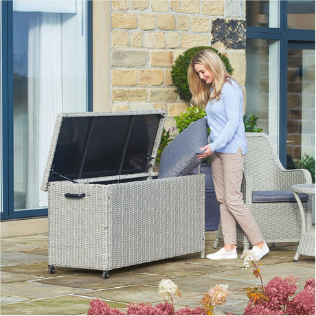 Woman on a patio lifting a cushion from an open Stone Grey Small Cushion Box, a rectangular synthetic rattan garden storage box on castors, beside matching outdoor armchairs.
