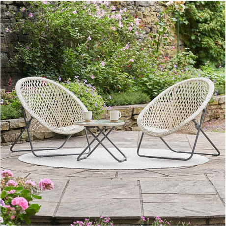 Tobs cream foldable lounger set with two round woven chairs and a matching glass-top side table, shown on a garden patio surrounded by greenery and flowers.
