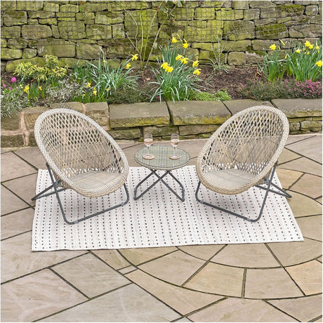 Tobs grey woven lounger chairs with a small glass-top side table on a patterned rug, set on a patio beside a stone wall and flowering border.