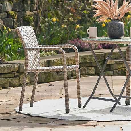 Tobs grey synthetic rattan stacking garden chair with woven aluminium frame and armrests, shown on a patio beside a folding table with mug and vase.