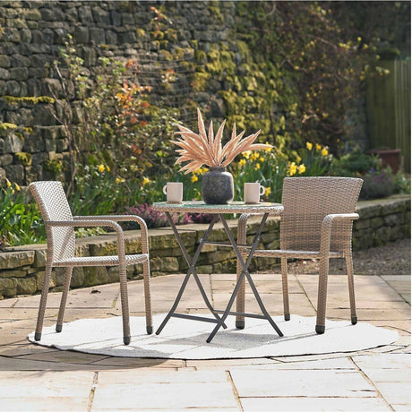Tobs Outdoor Grey Synthetic Rattan Stackable Bistro Set with round glass-top table and two chairs, shown on a garden patio with stone walls and flowers.