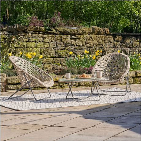 Tobs grey woven foldable loungers with a glass-top oval table, set on a patio rug in a garden with a stone wall and daffodils.