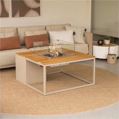Cosi Sandy Beige & Teak Gas Fire Pit Table, 100cm, with square teak top, beige metal base, glass wind guard, and lit burner, shown in a living room beside a sofa.