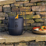 Cosi Basket Black Gas Flame Lantern, 31cm, with wire cage design and lit flame, shown outdoors on a stone ledge against a rustic stone wall.