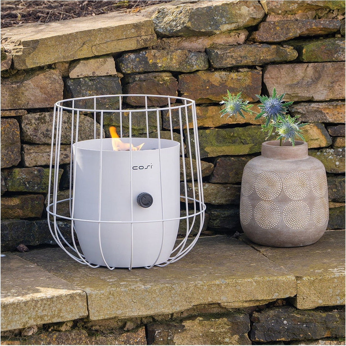 Cosi Basket White Gas Flame Lantern, 31cm, with wire cage frame and lit flame, shown on stone steps beside a decorative vase, outdoor garden setting.