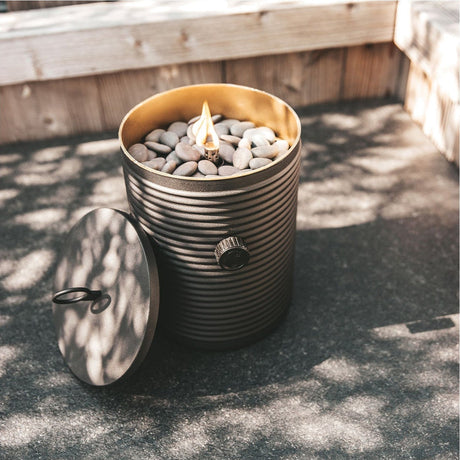 Cosi Iconic Black Gas Flame Lantern, 25cm, ridged steel cylinder body with lit flame amid pebbles, removable lid and control dial, shown outdoors on a patio.
